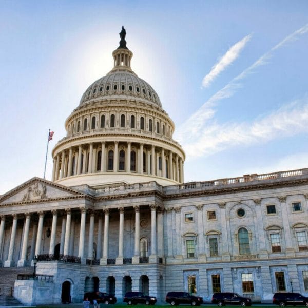 The U.S. Capitol building backlit by morning sunlight, with a blue sky and light, thin lines of clouds emanating from behind the rotunda.