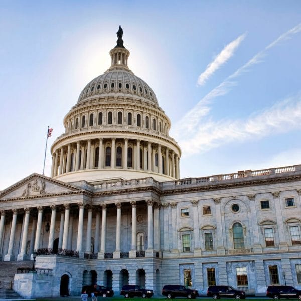 The U.S. Capitol building backlit by morning sunlight, with a blue sky and light, thin lines of clouds emanating from behind the rotunda.