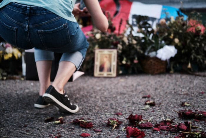 A woman in denim capris and black and white sneakers visits a memorial at the site of the shooting at Walmart in El Paso Texas. Out of focus beyond the woman are flowers, signs, and framed photos.